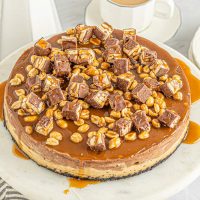 snickers cheesecake with bits of candy bar on top covered in caramel